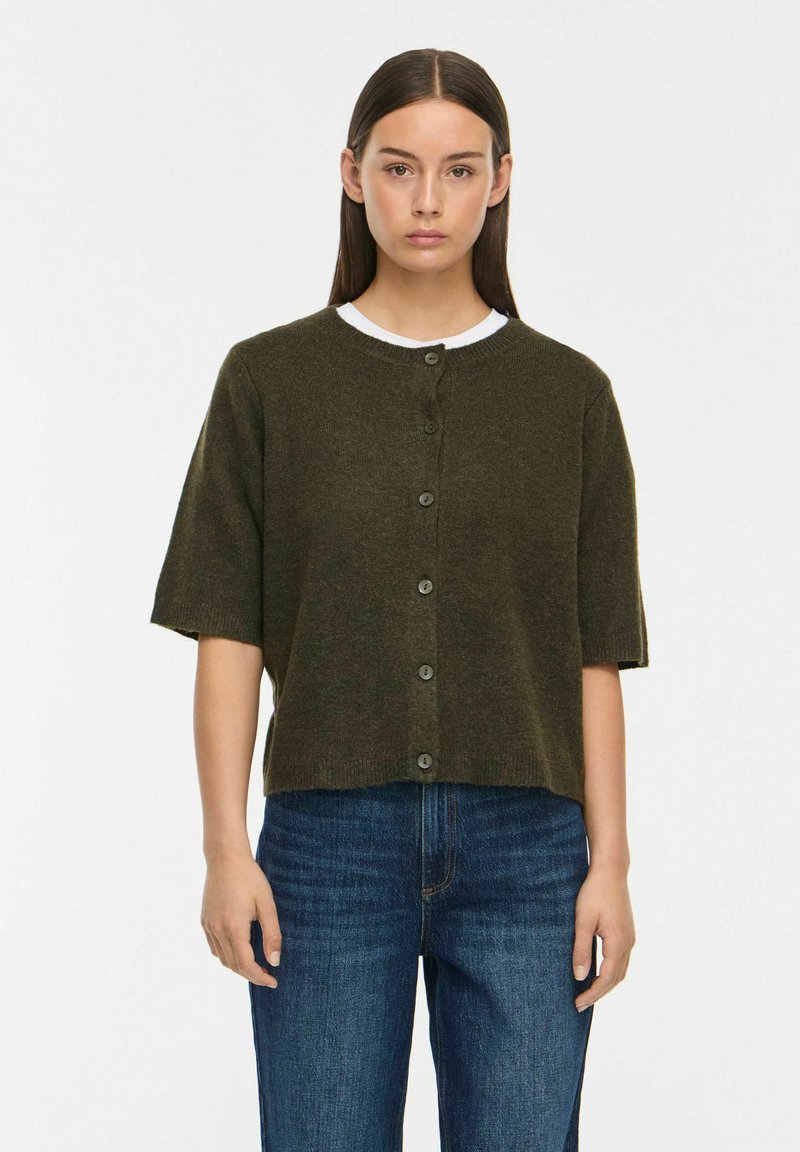 Femme aux cheveux bruns et lisses portant un cardigan court vert foncé boutonné sur une chemise blanche et un jean bleu, debout devant un fond blanc.