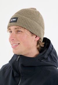 Nuori mies, jolla on päässään beigen värinen neulepipo, jossa on "SLOPE"-etiketti, sekä musta vetoketjullinen takki korkealla kauluksella ja säädettävällä hupulla.