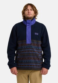 Fleece pullover met een marineblauwe basis, een paarse drukknoopkraag en een patroon in bruin en blauw op de onderste sectie. Bevat een buidelzak aan de voorkant.