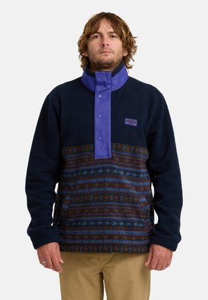 Fleece pullover met een marineblauwe basis, een paarse drukknoopkraag en een patroon in bruin en blauw op de onderste sectie. Bevat een buidelzak aan de voorkant.