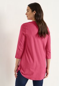 Vrouw met lang bruin haar draagt een felroze top met driekwart mouwen en donkerblauwe spijkerbroek, kijkt weg tegen een effen achtergrond.