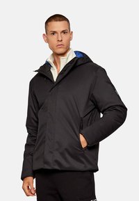 Giacca nera isolata con cappuccio, con accenti blu, due tasche laterali e chiusura con zip. Realizzata in materiale resistente e impermeabile.