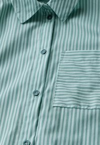 Turkoosi raidallinen nappikauluspaita, jossa on pehmeä rakenne. Paidassa on terävä kaulus, siniset napit ja vasemmassa rinnassa vaakaraidallinen rintatasku.