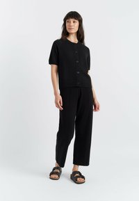 Schwarze kurze Strickjacke mit kurzen Ärmeln und sieben vorderen Knöpfen, kombiniert mit schwarzen weiten Hosen und schwarzen Sandalen. Durchgehend glatte Textur.