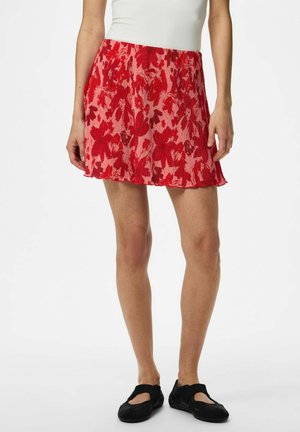 Femme portant une mini-jupe plissée rouge à motifs floraux, un haut blanc et des chaussures plates Mary Jane noires, debout sur un fond blanc.