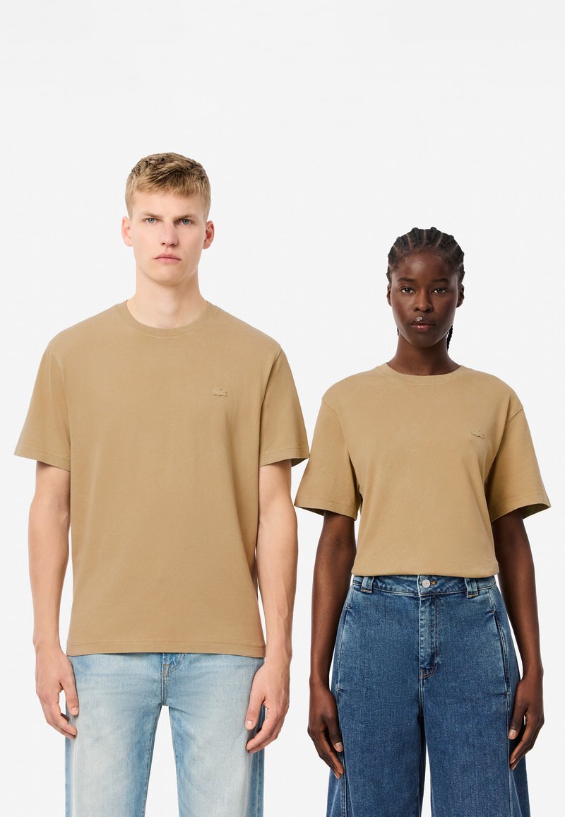 Zwei Personen tragen lässige, kurzärmlige T-Shirts in einem hellbraunen Farbton mit einem kleinen Logo. Der männliche Model trägt ein gerades Shirt, während das weibliche Model einen leicht oversized Schnitt hat.