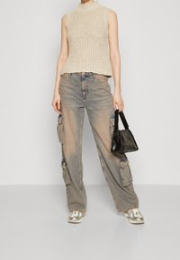 Häkelndes cremefarbenes ärmelloses Oberteil, lockere Passform, strukturiert. Kombiniert mit hellblauen Cargo-Jeans mit Taschen und einer schwarzen Handtasche. Silberne Sneakers.
