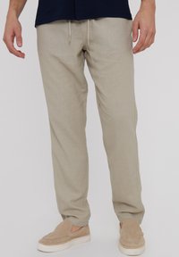 Beige Leinenhosen mit lässiger Passform, ausgestattet mit einem elastischen Kordelzugbund und Seitentaschen. Kombiniert mit hellbeigen Slippern.