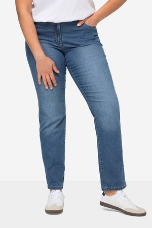 Person iført blå jeans med lige ben, hvide sneakers med gummisåler og en hvid skjorte, stående med den ene hånd i lommen.