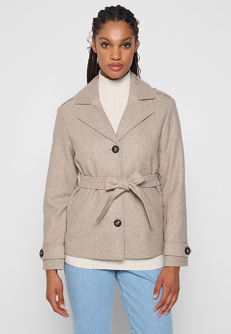 Donna che indossa un cappotto di lana beige con cintura sopra un maglione a collo alto bianco e jeans azzurri chiari, in piedi su uno sfondo neutro.