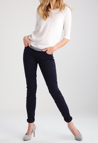 Weißes Hemd mit drei Viertel-Ärmeln und schwarz-weiß gestreifter Besatz, kombiniert mit körpernahen marineblauen Hosen und grauen Absätzen. Glattes Material, zeitgemäße Passform.