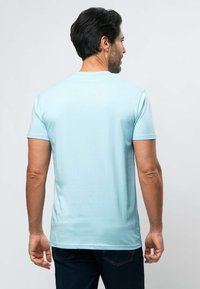 Vyras, stovintis nugara į priekį, dėvi šviesiai mėlyną trumpomis rankovėmis marškinėlį ir tamsiai mėlynas džinsines kelnes, stovintis priešais paprastą šviesią foną.