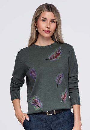 Femme aux longs cheveux blonds portant un pull vert foncé avec des motifs colorés de plumes et un jean bleu sur un fond uni.