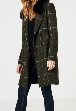 Vrouw draagt een knie-lange donkergroene geruite jas met een zwarte bontkraag, gecombineerd met een zwart shirt en donkere skinny jeans.