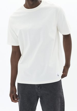 T-shirt blanc à manches courtes en coton avec un col rond. Dispose d'une petite étiquette "73" sur la couture en bas à gauche.