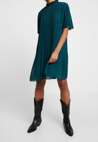 Personne portant une robe plissée sarcelle longueur genou avec un col à volants et des bottes de cowboy noires mi-mollet, debout devant un fond uni.