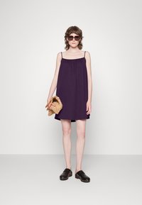 Vestido roxo sem mangas, com um corte solto e decote franzido, emparelhado com tamancos pretos e uma bolsa de palha em tom bege.