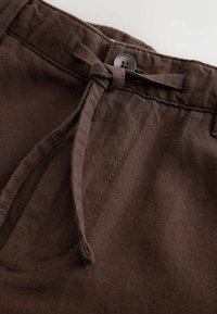 Gros plan sur la ceinture d’un pantalon marron avec un cordon en tissu noué en nœud et une fermeture par bouton noir à quatre trous.