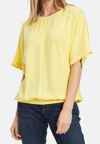 Żółta bluzka z krótkim rękawem, luźny krój i okrągły dekolt. Teksturowana tkanina z falbaną na dole, zestawiona z ciemnoniebieskimi dżinsami.