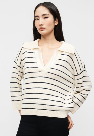 Νεαρή γυναίκα με μακριά σκούρα μαλλιά, ντυμένη με κρεμ πουλόβερ με μαύρες οριζόντιες ρίγες και βαθύ V-λαιμόκοψη, ποζάροντας μπροστά σε απλό φόντο.