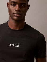 Camiseta preta de manga curta com textura em malha nos ombros, apresentando um logótipo branco "CALVIN KLEIN" na frente. Acabamento de tecido suave.
