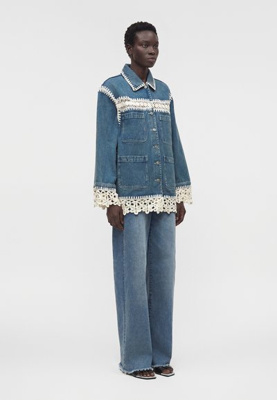 Γυναίκα με μπλε τζιν μπουφάν και ευρύχωρο τζιν παντελόνι, και τα δύο με λευκή δαντέλα, στέκεται μπροστά σε ένα απλό φωτεινό φόντο.