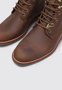 Botas de cuero marrón hasta el tobillo con puntera redonda, diseño de cordones, ojales dorados, acentos blancos cosidos y suela de goma texturizada.