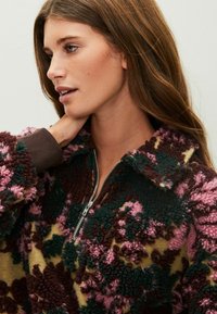 Fleecový pulóver so zapínaním na zips, kvetinovým vzorom v ružovej, zelenej a burgundskej farbe, s textúrovaným, plyšovým povrchom a hnedými rebrovanými manžetami.