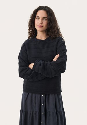 Mornarsko modra pletena jopica s vodoravnimi črtami, rebrasto teksturo, okroglo ovratnico in gumbnimi detajli na ramenih. Oblečena čez temno večerno obleko.