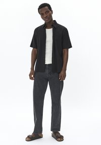 Chemise noire à manches courtes avec boutons, débardeur écru, pantalon en denim gris, sandales noires. Look décontracté avec un design relax et un matériel minimal.