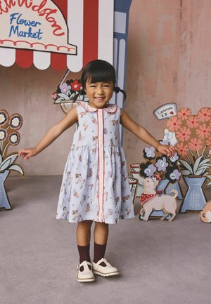 Fată zâmbitoare cu cozi, purtând o rochie deschisă cu imprimeuri cu animale, stând cu brațele întinse în fața unui fundal colorat cu o piață de flori.