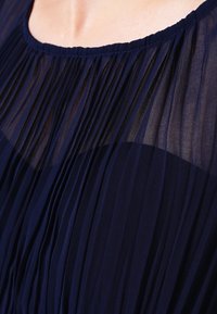 Närbild av en person som bär en genomskinlig, plisserad marinblå topp med en synlig rund halsringning och en delikat tygstruktur.