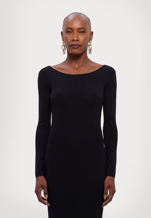 Abito lungo a maniche lunghe di colore nero con un ampio scollo a barca. La texture liscia e la forma aderente valorizzano la silhouette. Grandi orecchini a cerchio metallici.