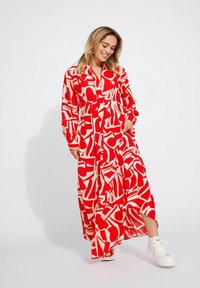 Robe maxi à motifs géométriques rouges et crème, manches longues, fermeture à boutons sur le devant, avec fentes latérales, associée à des baskets blanches.
