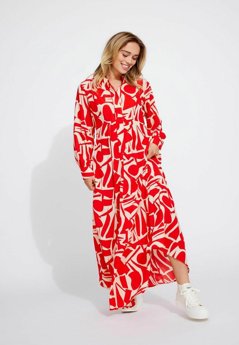 Robe maxi à motifs géométriques rouges et crème, manches longues, fermeture à boutons sur le devant, avec fentes latérales, associée à des baskets blanches.