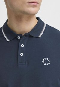 Marineblå poloskjorte med teksturert stoff, hvite detaljer på kragen og tre knapper. Har en rund logo på venstre brystområde.