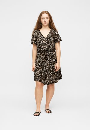 Kobieta o długich włosach stoi przodem, ubrana w sukienkę z krótkim rękawem w panterkę i czarne sandały na jednolitym białym tle.