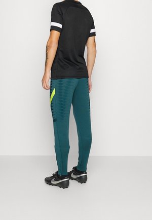 Henkilö, joka pukeutuu mustavalkoiseen lyhythihaiseen paitaan, petroolinsinisiin kuvioituihin urheiluhousuihin neonyksityiskohdilla ja mustiin Nike-piikkareihin, seisoo valkoista seinää vasten.