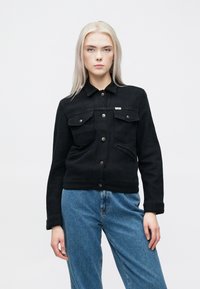 Crna traper jakna s kragnom, dva džepa na prsima s preklopima i metalnim kopčama. Prilagođen kroj, teksturirana tkanina.