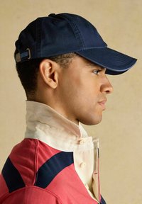 Mies, jolla on päässään tummansininen lippalakki ja yllään punainen takki, jonka kaulus on kermaan vivahtava ja korkea, katsoo oikealle beigeä taustaa vasten.