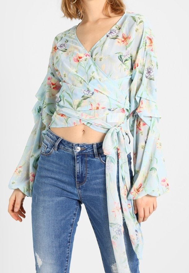 Světlomodrá blůza s květinovým vzorem, s volány na dlouhých rukávech a detaily vázání v pase, spárovaná s vybledlými modrými džíny.