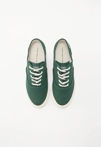 Grønne lerretssneakers med hvite snørebånd, rund tå og gummisåle. Har logo på tungen og kontrasterende søm langs sidene.