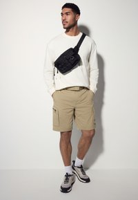 Black crossbody bag with textured pattern, beige cargo shorts, white long-sleeve shirt, and multicolored athletic shoes with black accents.