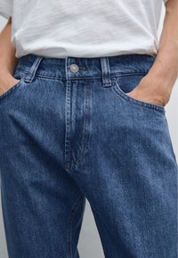 Pantalon en denim bleu foncé avec une texture délavée. Présente un design classique à cinq poches, une fermeture par bouton en métal et des coutures visibles.