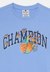 T-shirt en coton bleu clair avec "CHAMPION" en lettres foncées, accompagné de graphiques colorés de flammes et de basket-ball en dessous.