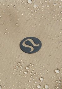 Beige Stoff mit Wassertropfen und einem runden schwarzen Logo in der Mitte. Das Material wirkt glatt mit einer dezenten Textur.