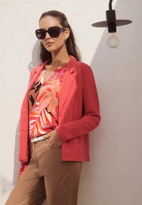 Korallenfarbener Blazer über einem mehrfarbigen Blumenoberteil, beige Hosen und einem weißen Gürtel. Mit dunklen quadratischen Sonnenbrillen accessorisiert. Texturierter Hintergrund.