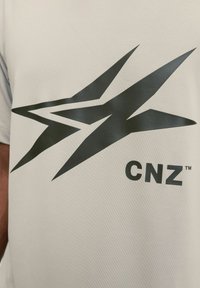Lys grå t-shirt med et iøjnefaldende sort grafisk design af tre vinklede former og teksten "CNZ" i fed sort skrift.