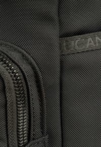 Borsa in tessuto nero con superficie strutturata, dotata di una cerniera prominente e di un'etichetta con il marchio integrata nel design.