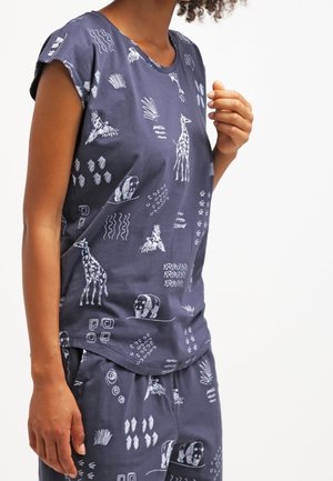 Persona con pijama azul oscuro de manga corta con estampados blancos de animales y formas geométricas, de pie sobre un fondo claro y liso.